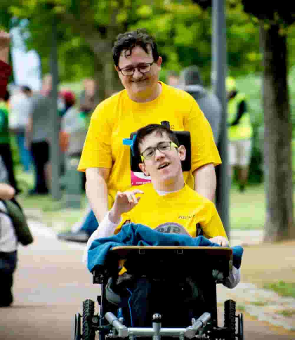 A man and woman in yellow shirts riding on the back of a wheelchair.